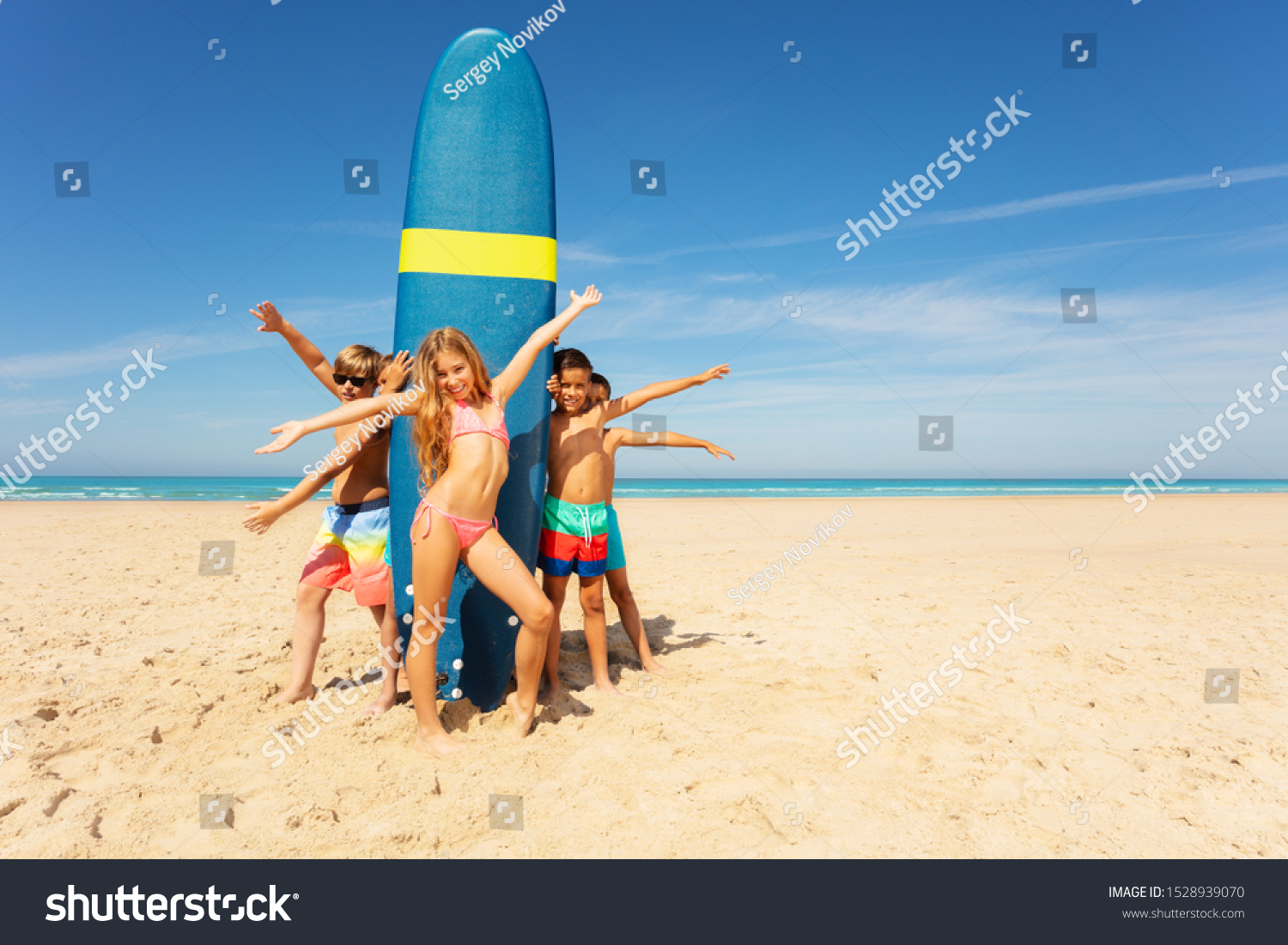 Group of kids stand waving hands near surfboard