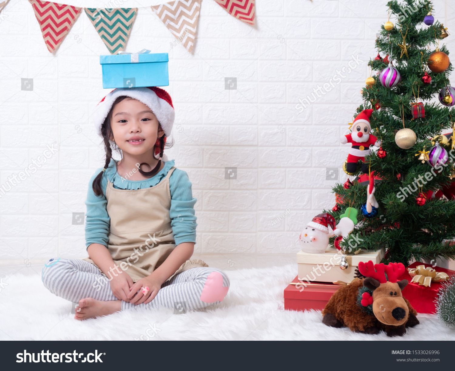 Asian little cute girl 6 years old sitting on carpet and put a gift box on her head in white room with the Christmas tree. Christmas celebration concept.