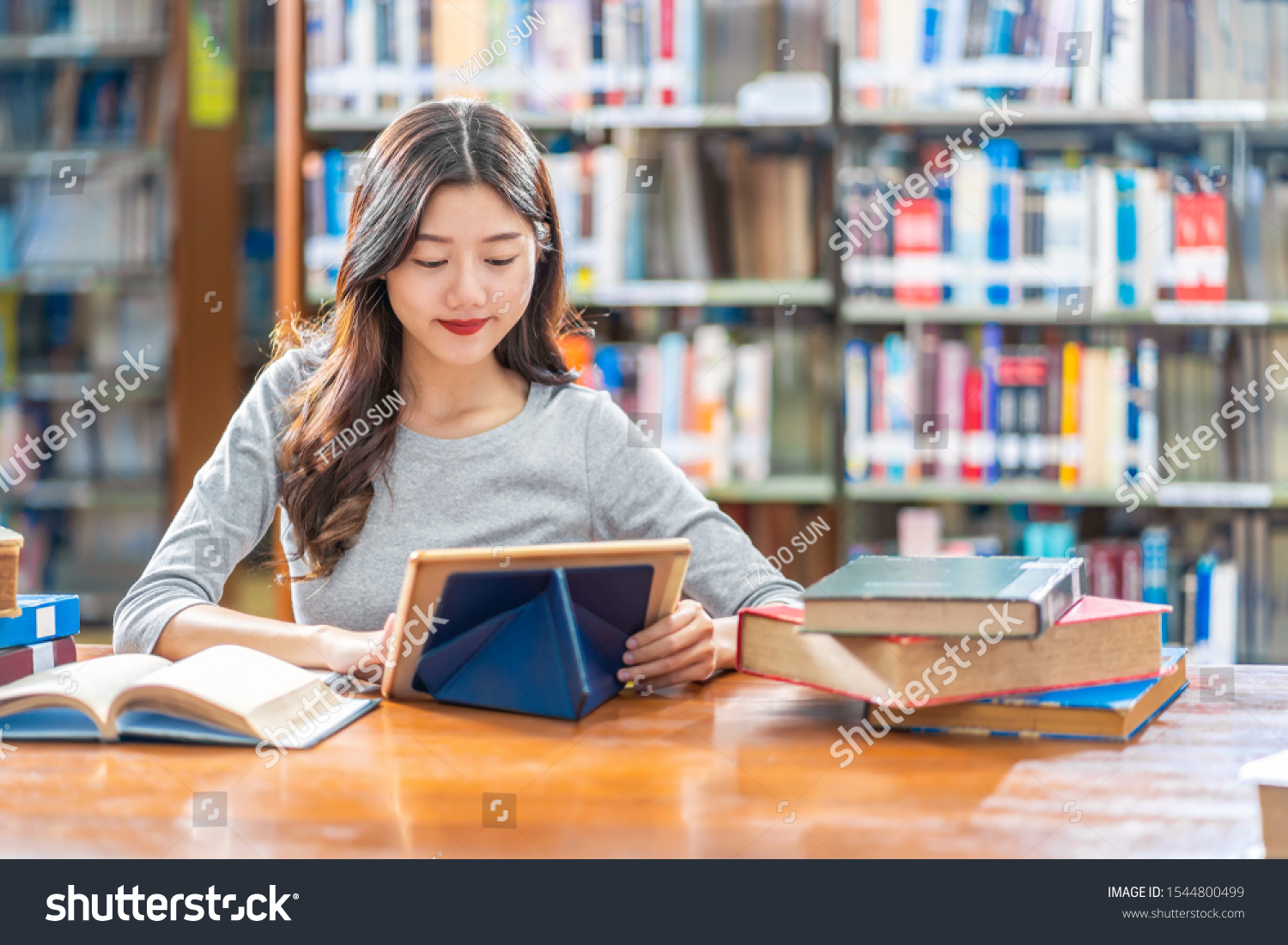 Asian young Student in casual suit doing homework and using technology teblet in library of university or colleage with various book and stationary over the book shelf background Back to school