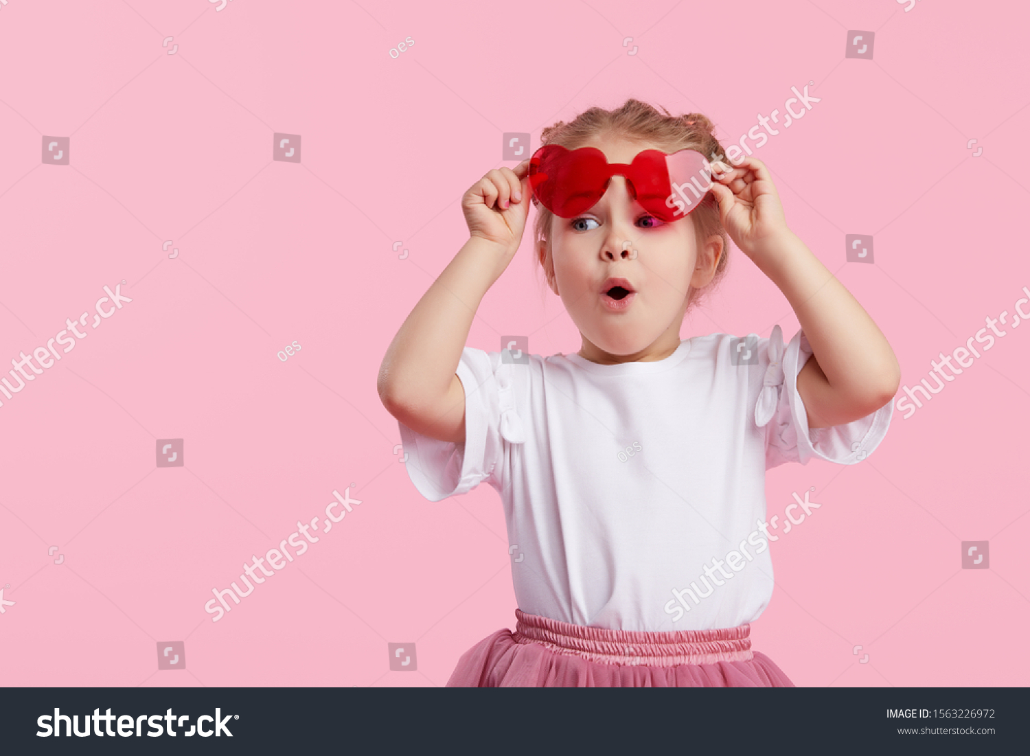 Portrait of surprised cute little toddler girl in the heart shape sunglasses. Child with open mouth having fun isolated over pink background. Looking at camera. Wow funny face