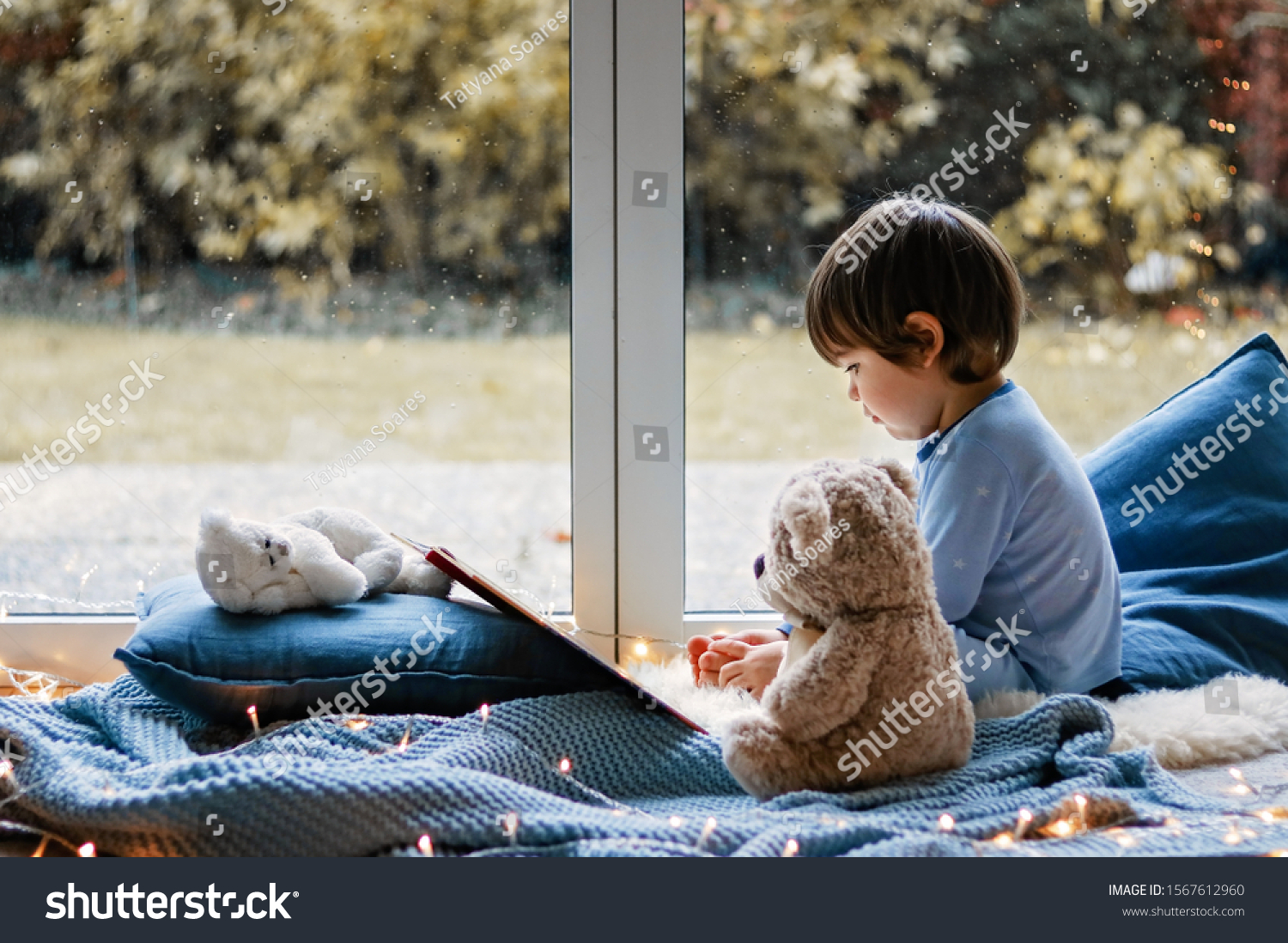 Cute little boy reading book with his teddy bear toy sitting cozy on pillows and knitted blanket near wet window with autumn garden at background. Cozy home. Winter holidays lifestyle.
