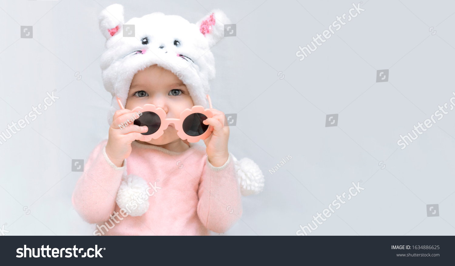little girl in fluffy hat holds sunglasses in pink dress on white gray background. Pastel natural colors. Fashion concept for kids children s style change of seasons. Banner copy space