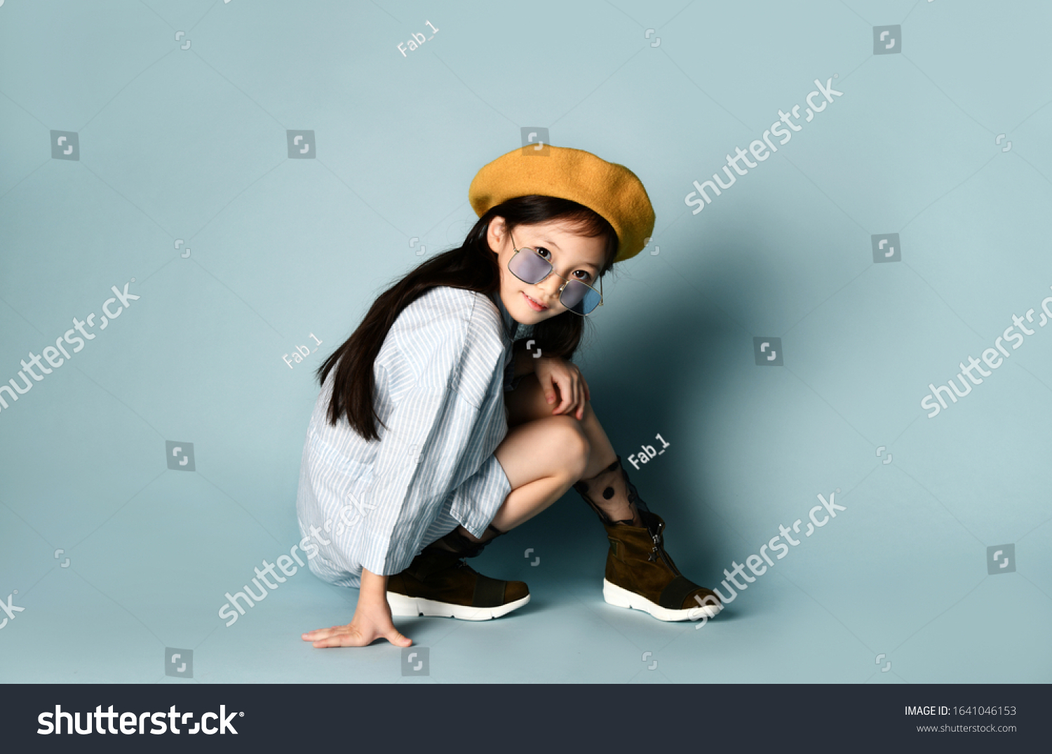 Little asian child in sunglasses  shirt dress  brown beret  boots. She smiling  posing cross-legged sitting on floor against blue background. Childhood  fashion  hipster style. Close up  copy space