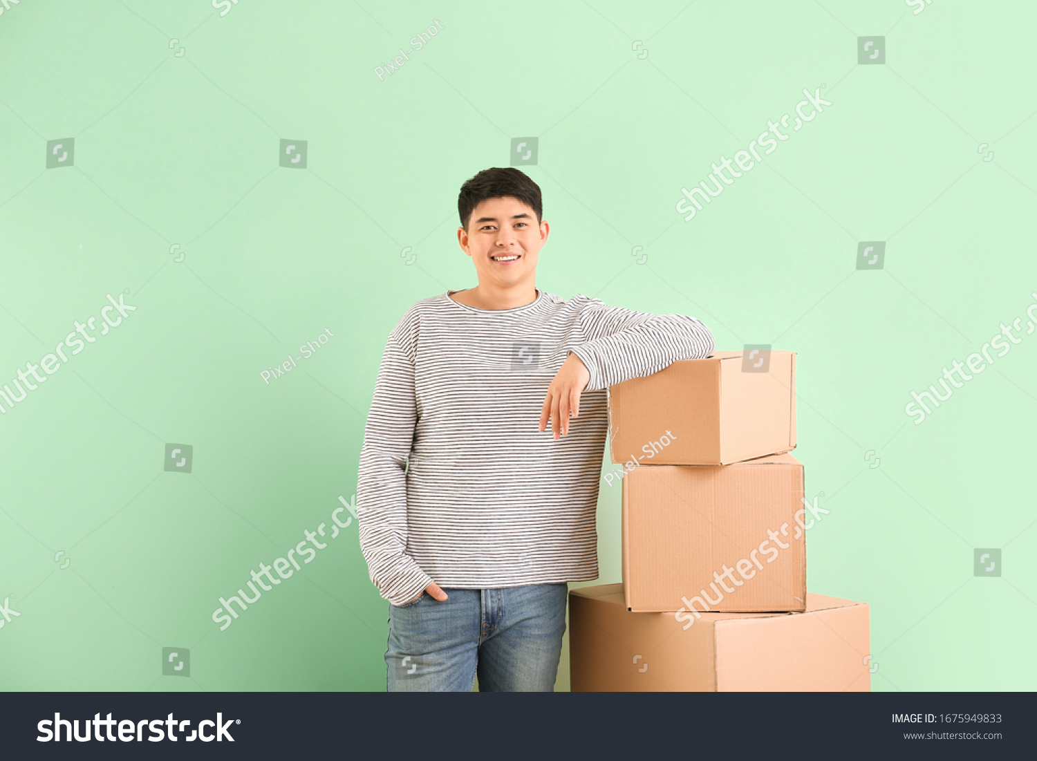 Asian man with moving boxes on color background