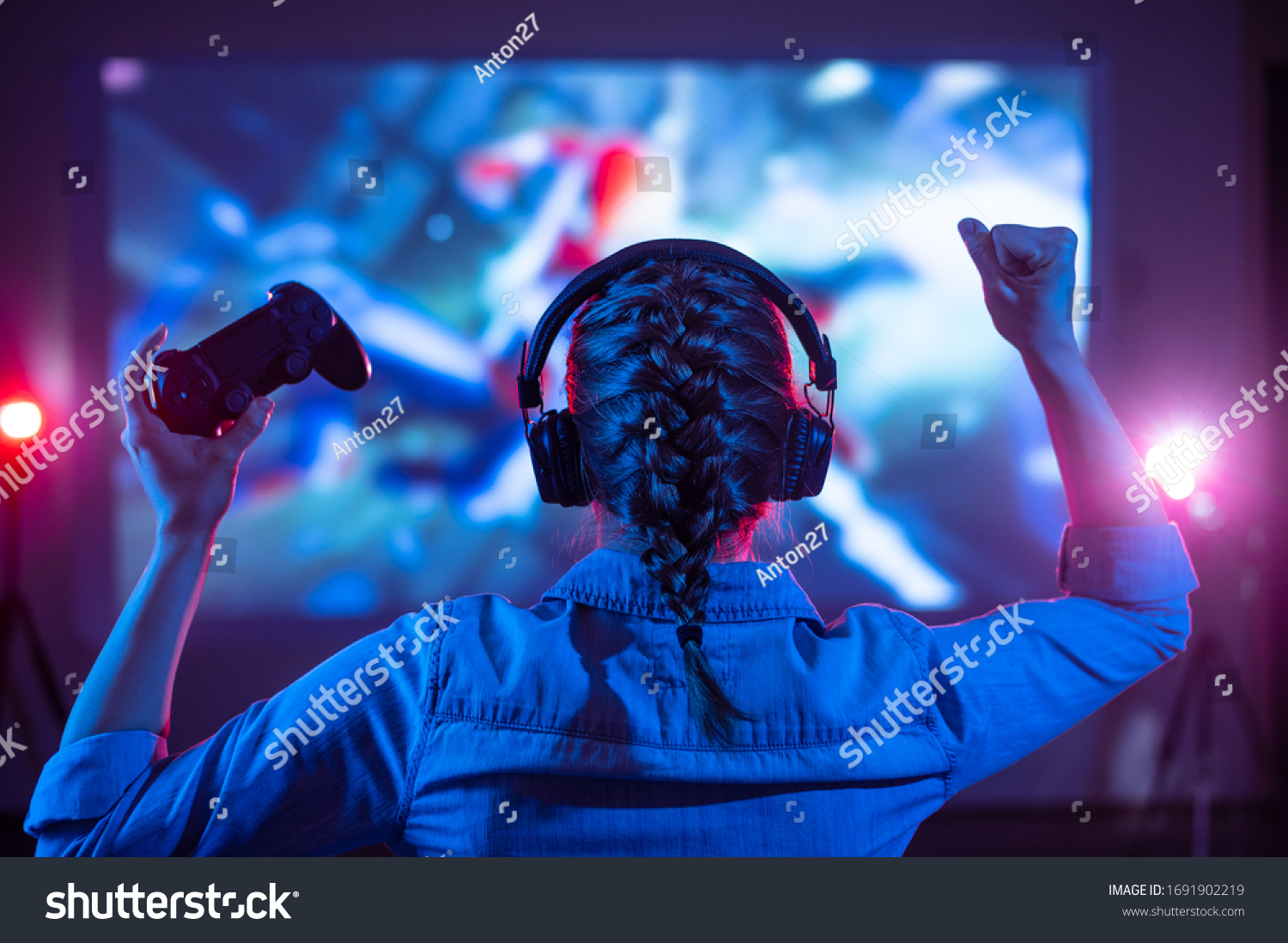 Girl in headphones plays a video game on the big TV screen. Gamer with a joystick. Online gaming with friends win prize. Fun entertainment. Teens play adventure games. Back view. Neon lighting