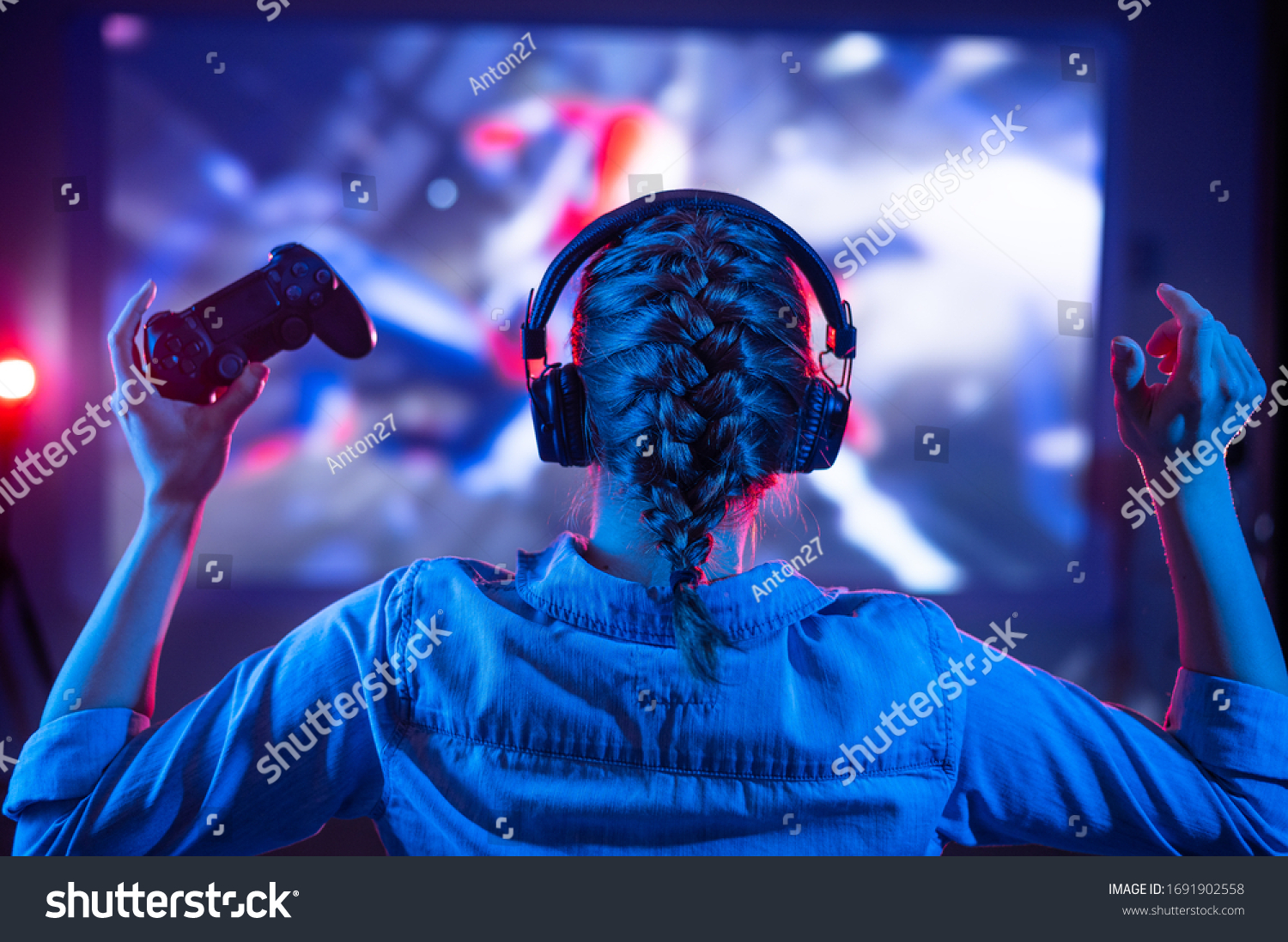 Young girl in headphones plays a video game on the TV in the dark room. Gamer with a joystick. Online gaming with friends competitions win. Fun entertainment. Teenagers play puzzles games. Back view