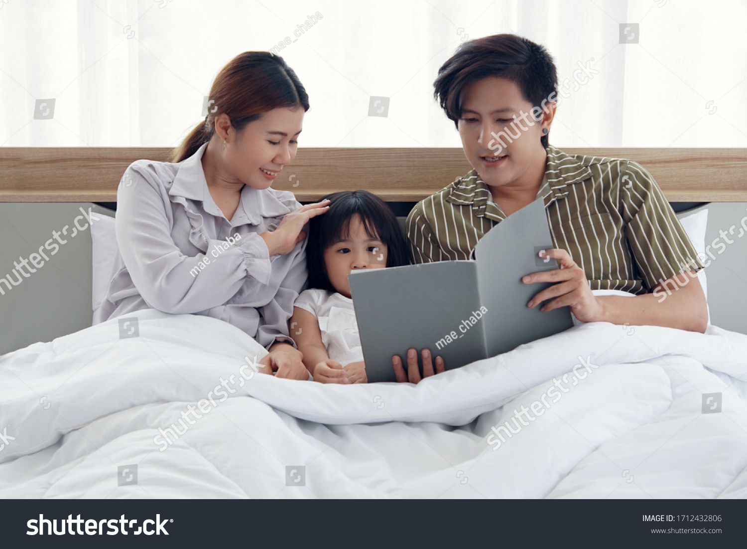 Happy asian family with a daughter lying sitting on the bed under the blanket The father is reading a book telling the stories to his daughter.the mother is pat her daughter’s head with love and care