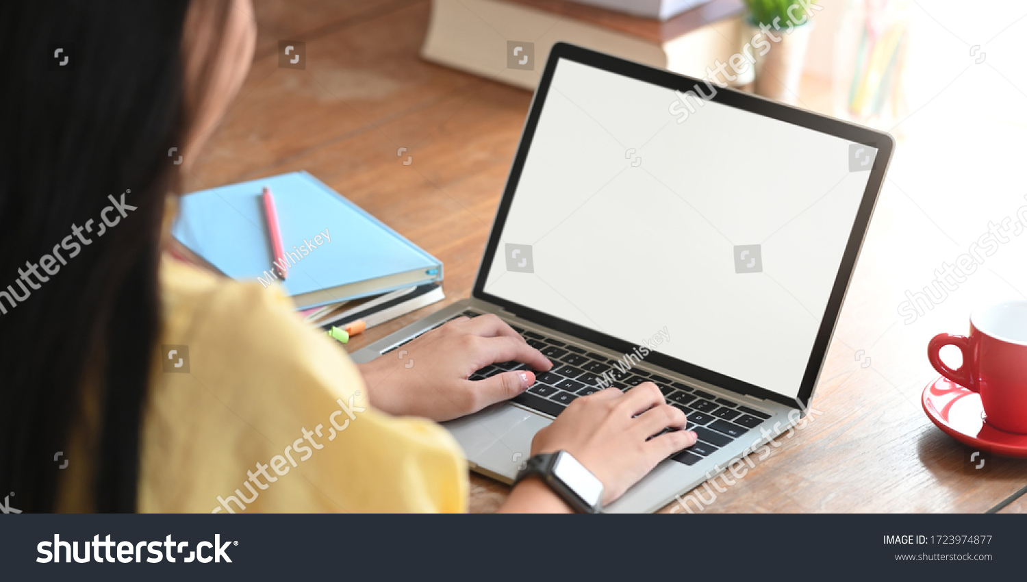 Behind shot of beautiful woman working as accountant while sitting and working with white blank screen computer laptop at the wooden working desk that surrounded by coffee cup and stack of books.
