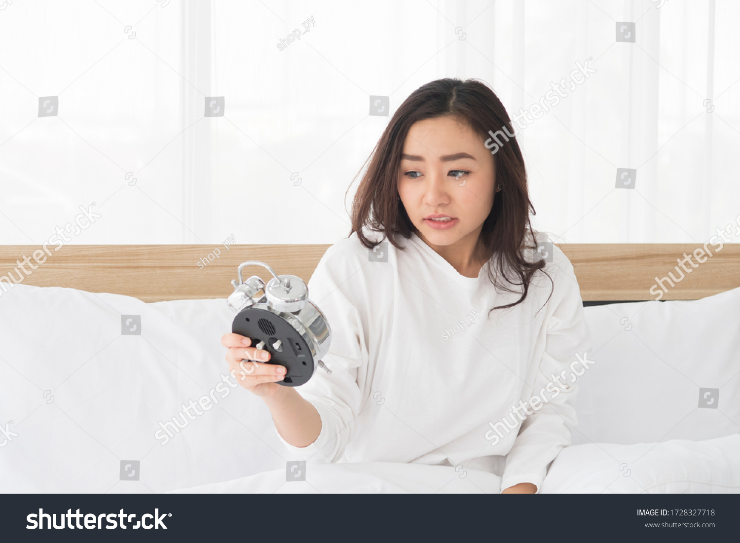 Portrait of Young Beautiful Asian bad moody woman shocked looking at the clock and sitting in white bed while wake up late in the morning.