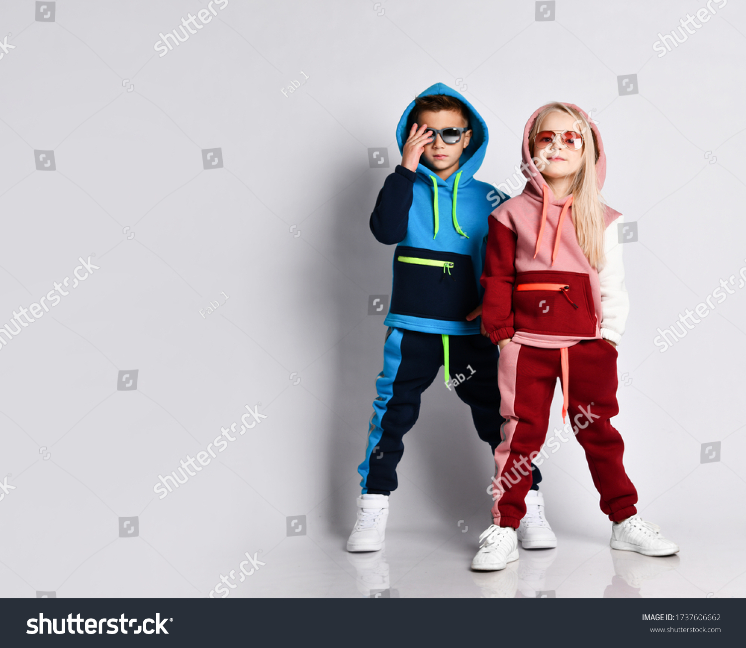 Little kids boy and girl in sunglasses and hoods colorful tracksuits sneakers. They posing isolated on white studio background. Childhood fashion advertising and sport. Full length copy space