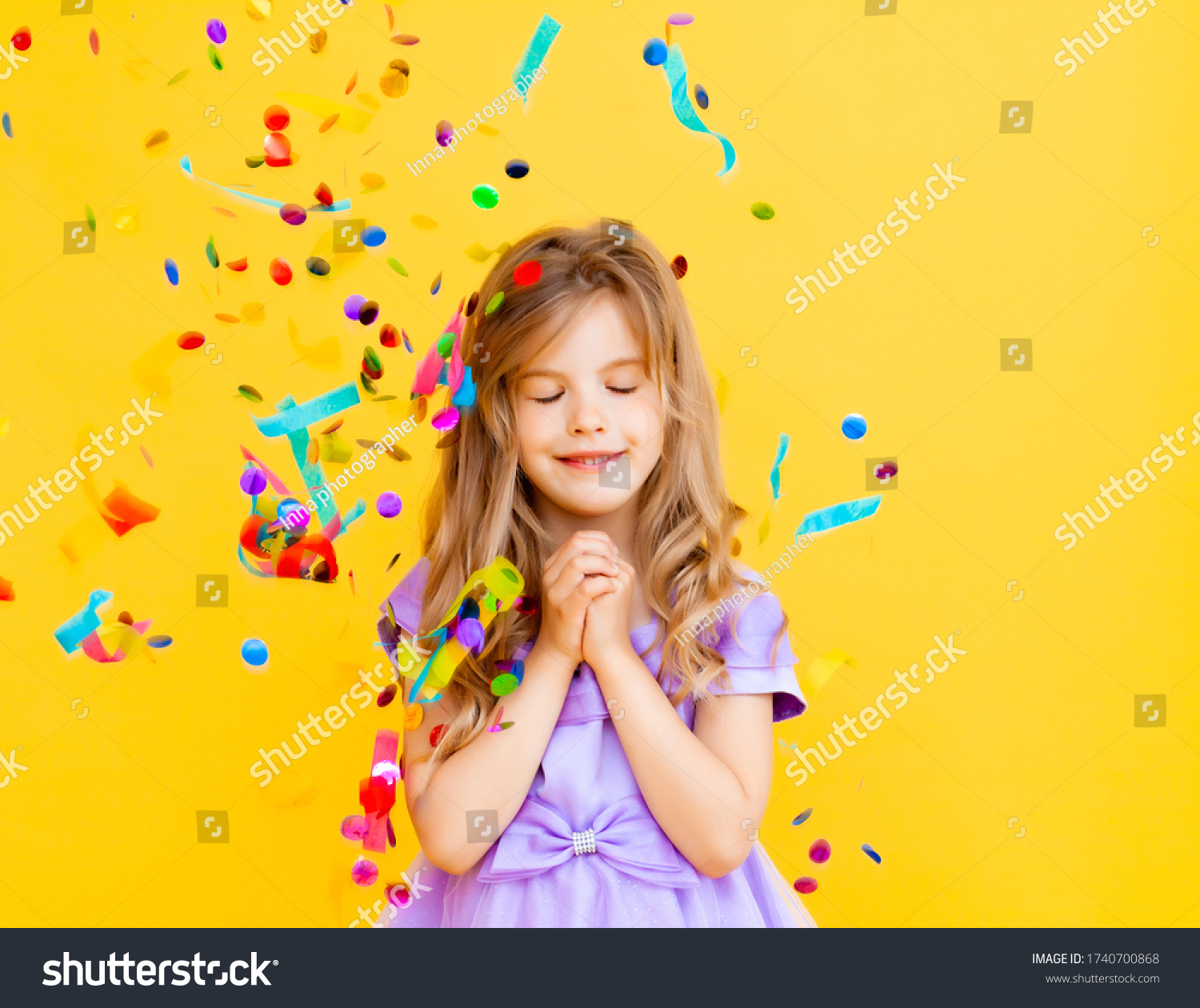happy little girl with blond hair and in a blue dress catches confetti on a yellow background holiday concept