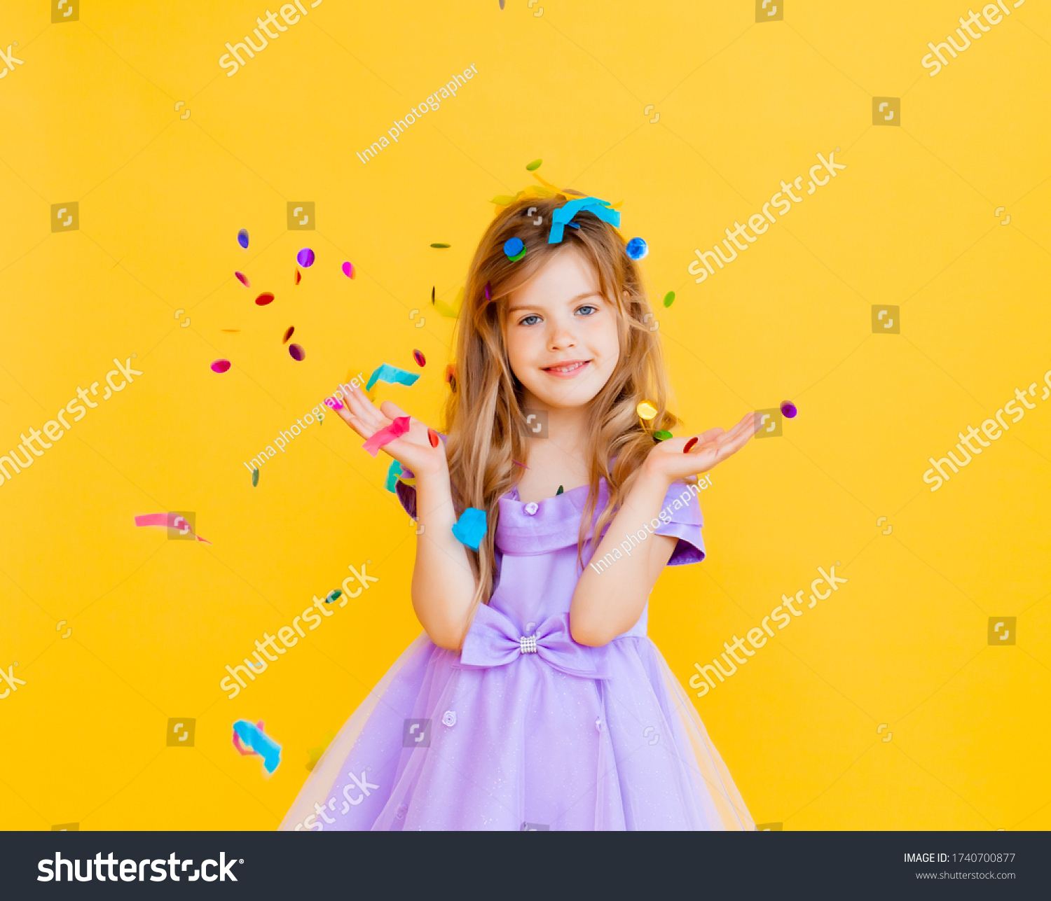 happy little girl with blond hair and in a blue dress catches confetti on a yellow background holiday concept