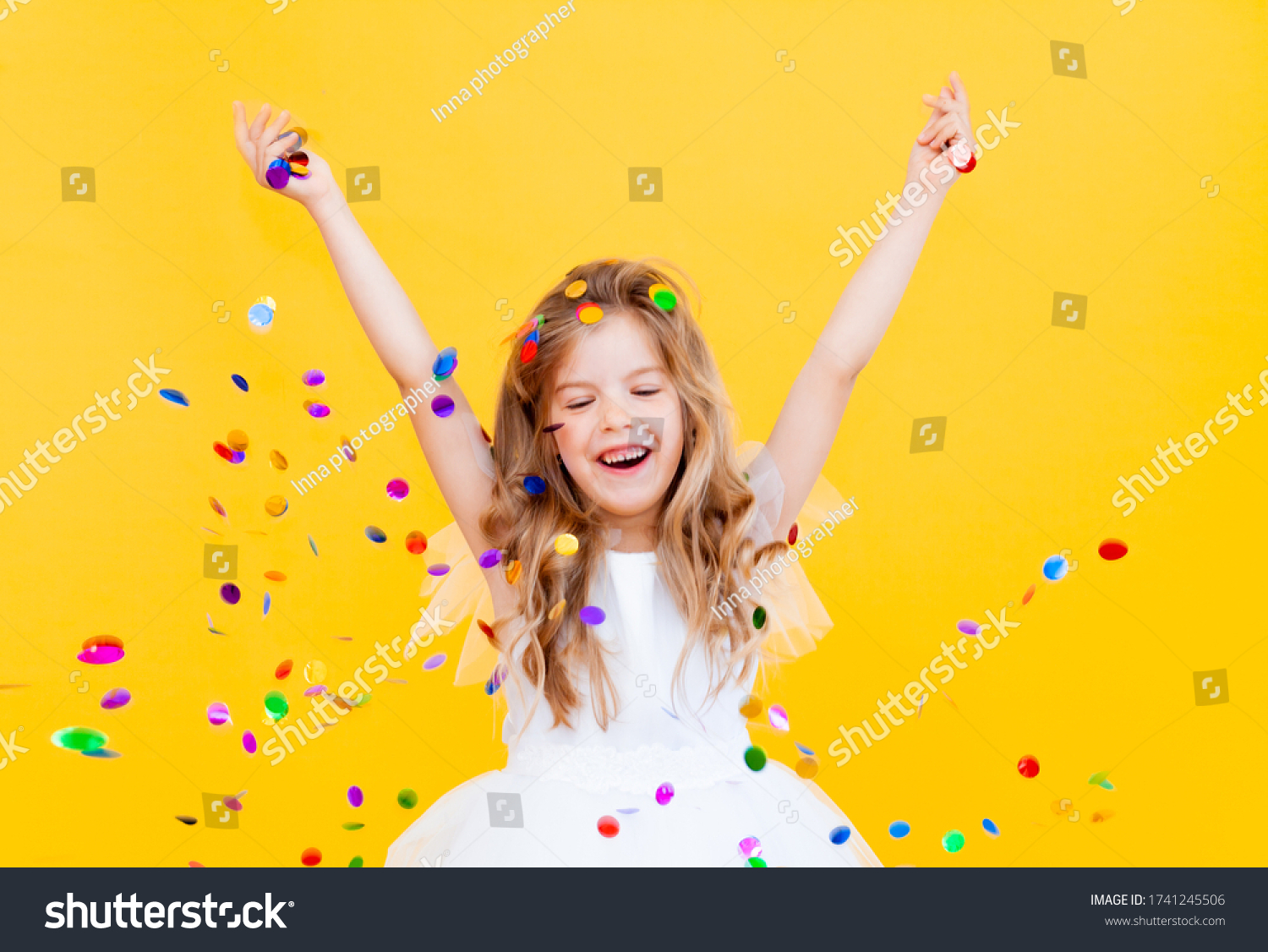 happy little girl with blond hair and in a white dress catches confetti on a yellow background holiday concept