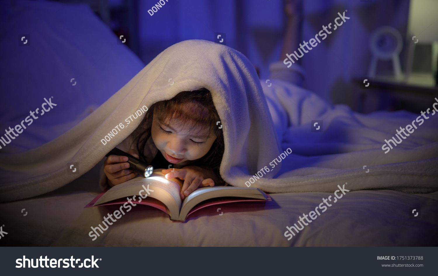 Child asian girl reading a book with flashlight under the blanket on bed in a dark bedroom at night before sleeping  Comfortable children at home concept