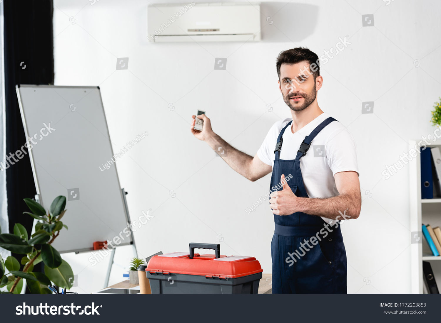 smiling worker holding remote controller and showing thumb up while standing near toolbox and air conditioner