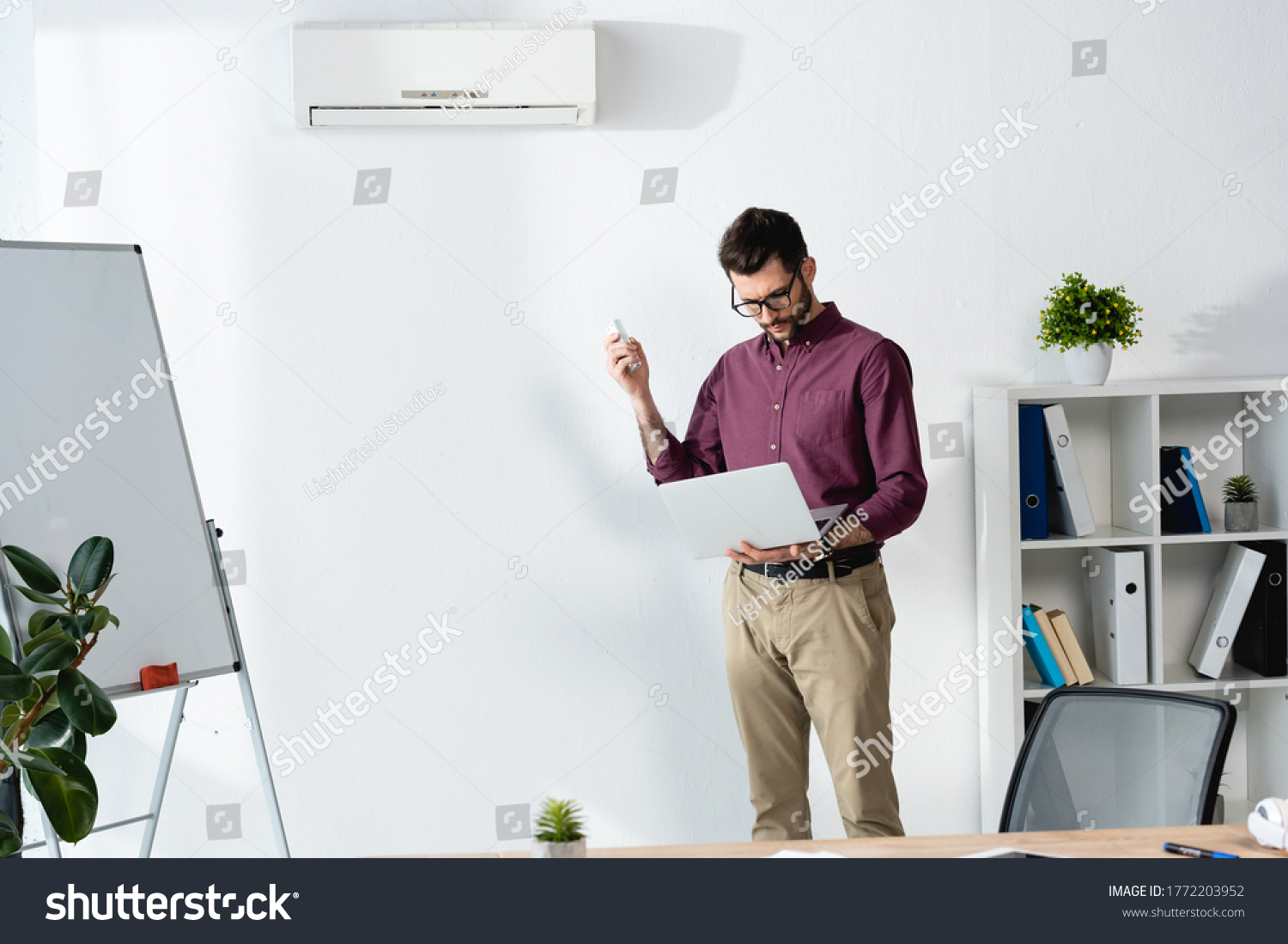handsome businessman holding laptop and switching on air conditioner with remote controller