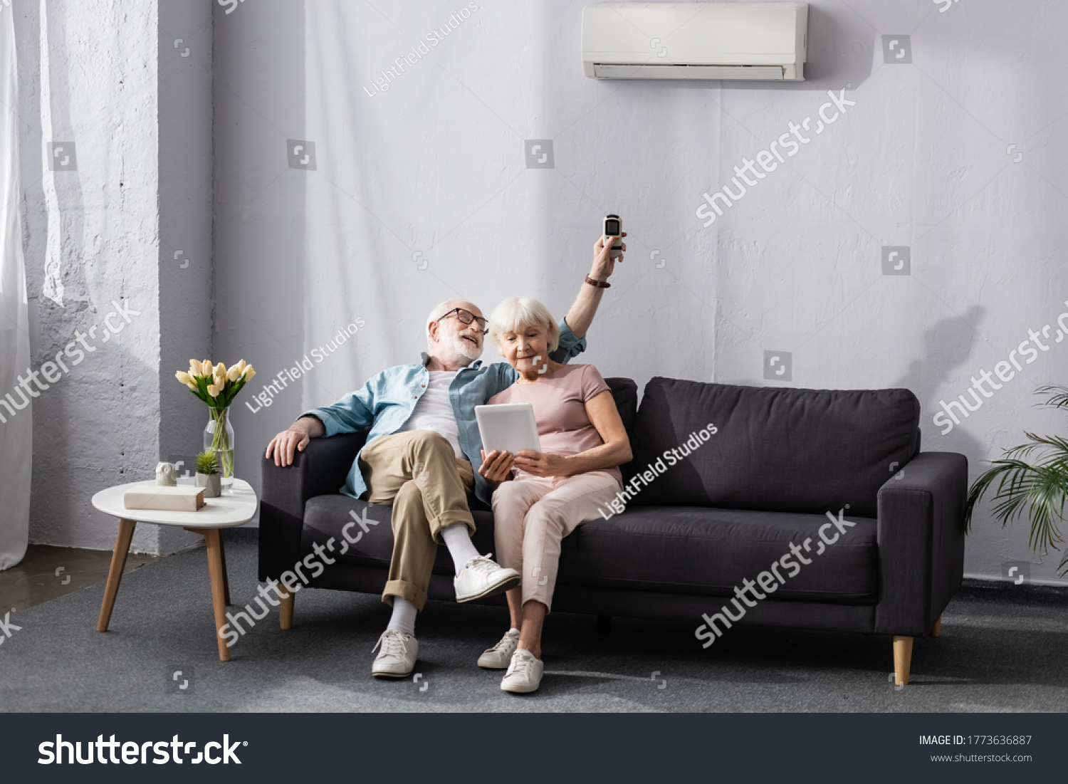 Senior woman using digital tablet near smiling husband switching air conditioner with remote controller