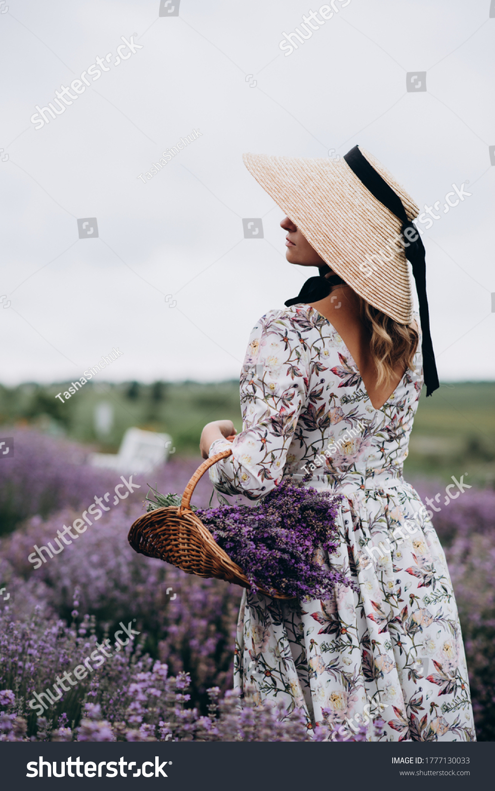 Young beautiful blonde woman in romantic dress straw hat and basket of flowers posing in lavender field back view. Soft selective focus.