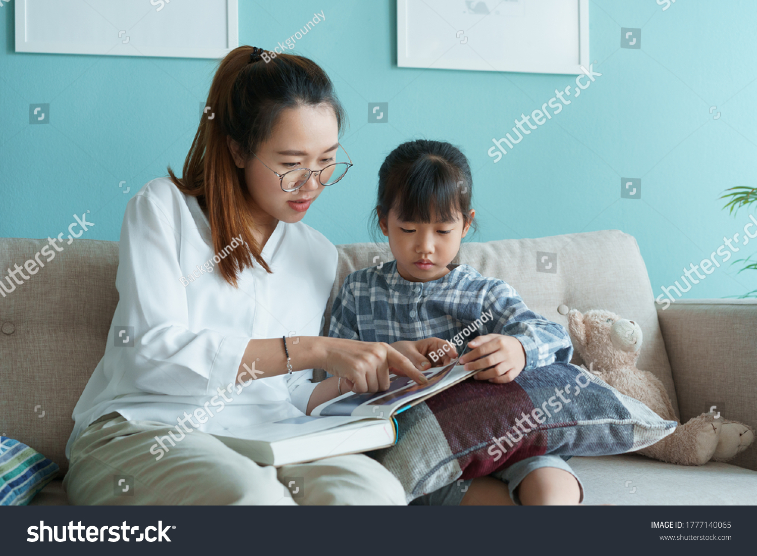 Asian family with mother and daughter reading book together while sitting on the sofa in the living room at home morning