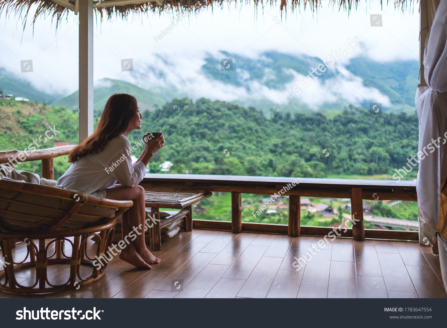 Portrait image of a beautiful asian woman holding and drinking hot coffee sitting on balcony and looking at mountains and green nature