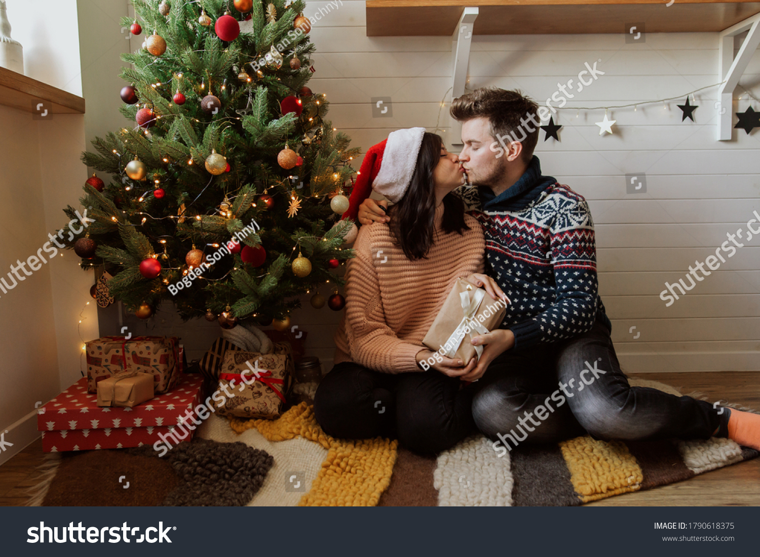 Stylish happy couple exchanging christmas gifts under christmas tree with lights. Young family kissing and holding present happy moment in festive modern room. Merry Christmas