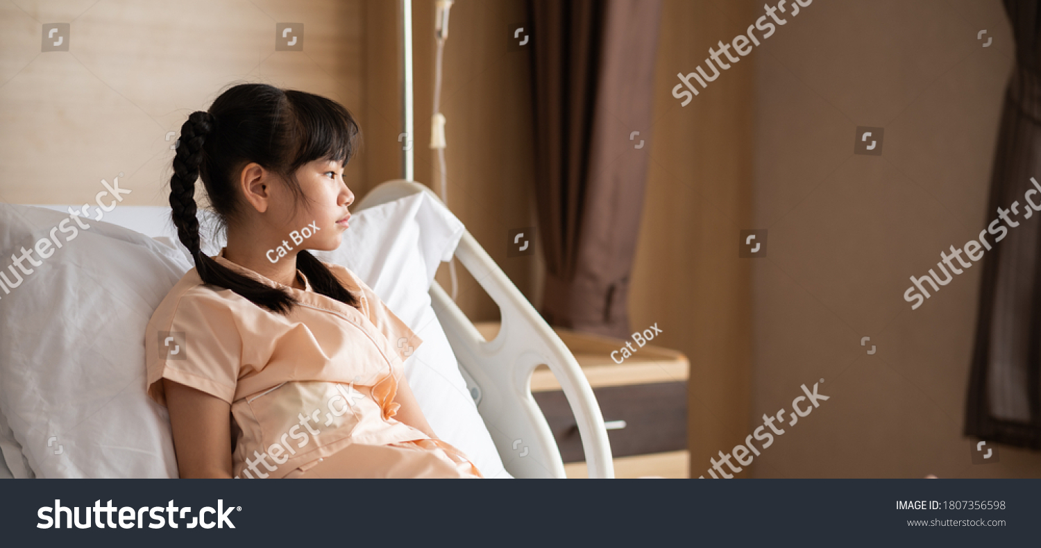 Sick Asian little girl sitting on hospital bed feeling sad and unhappy. Homesick girl looking hopefully outside and want to go home soon while being treated in the hospital. Banner ratio photo.