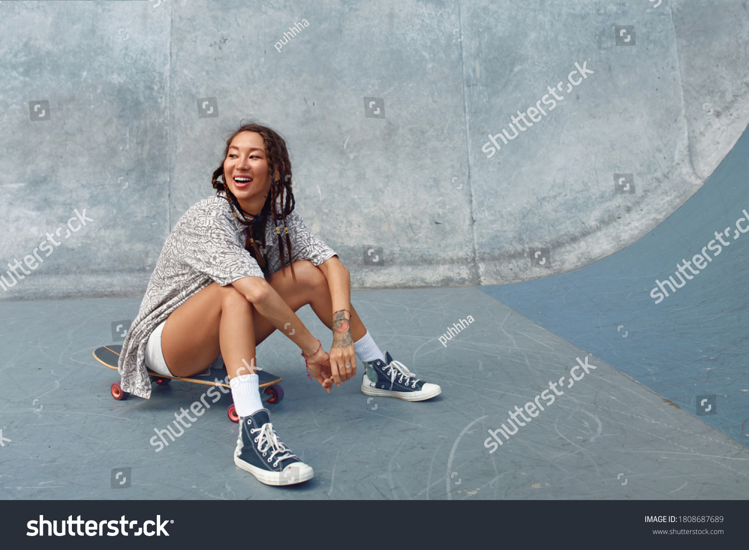 Portrait Of Skater Girl In Skatepark. Female Teenager In Casual Outfit Sitting On Skateboard Against Concrete Wall. Summer Skateboarding With Modern Sport Equipment As Part Of Active Lifestyle.