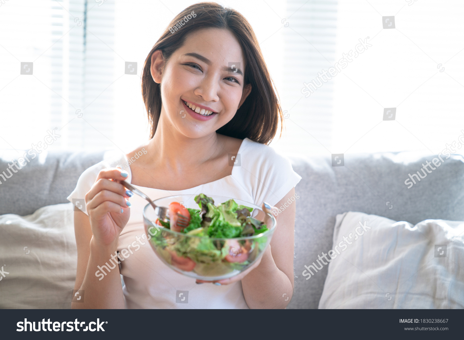 Young happy Asian woman eating healthy salad sitting on the sofa in living room.