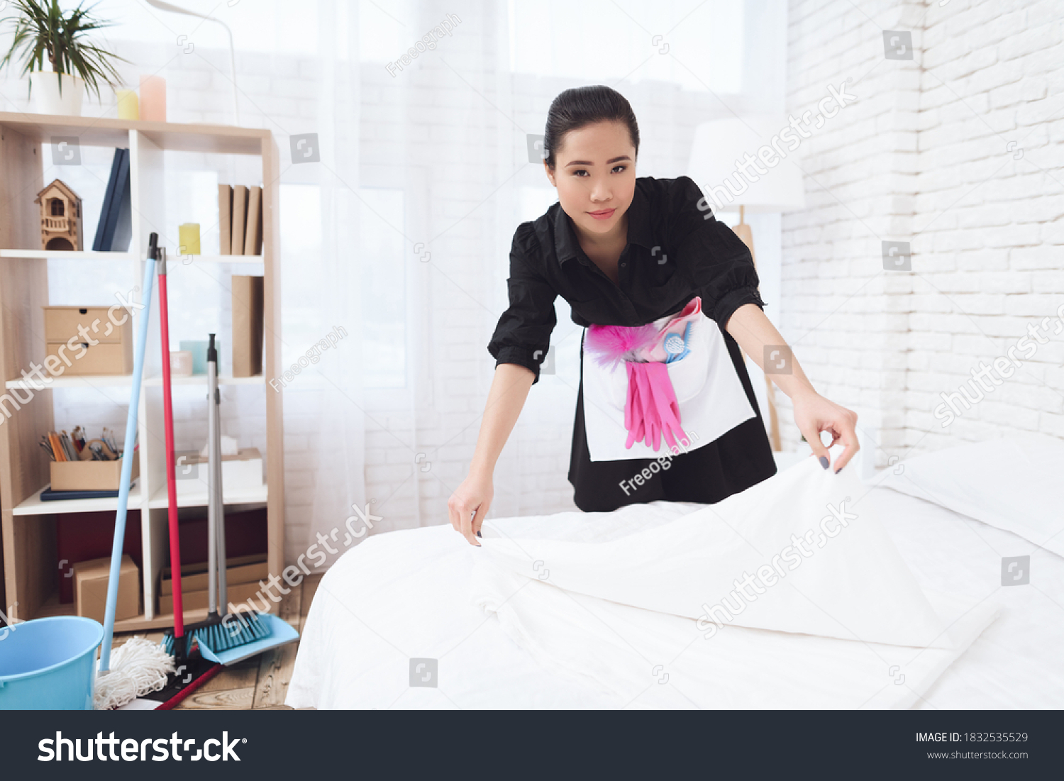Chinese housemaid woman is changing bed at house. House cleaning. Asian woman in special uniform looks at the camera while making a bed.