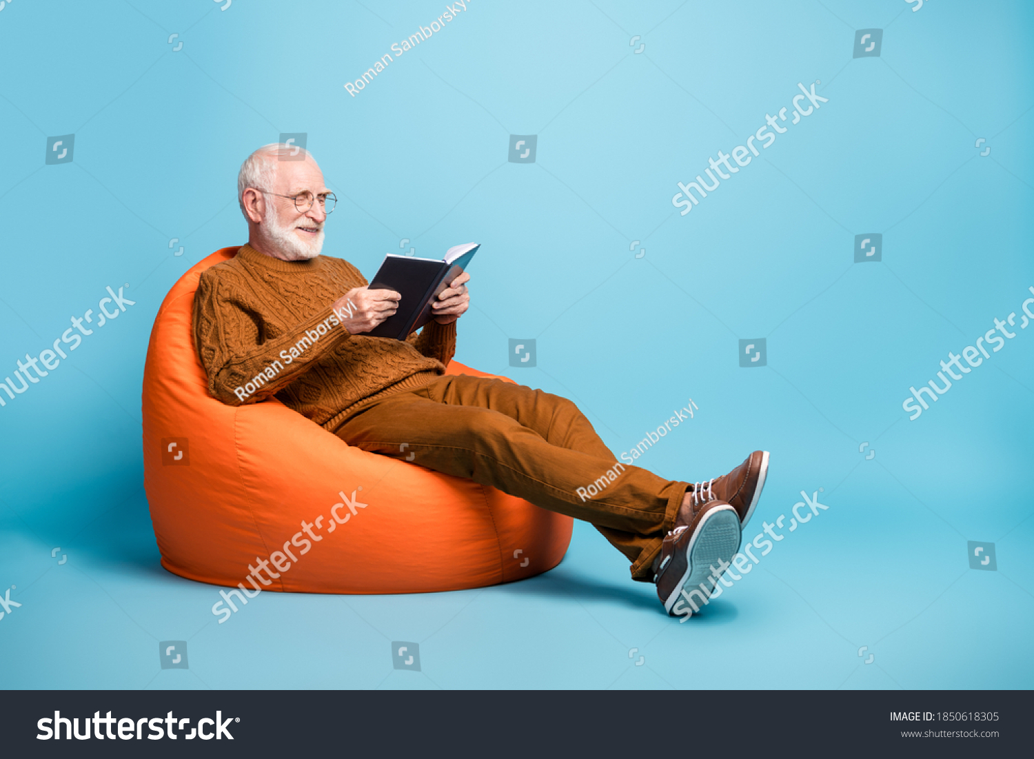 Portrait of his he nice attractive focused cheerful cheery wise smart clever bearded grey-haired man sitting in bag chair reading academic book isolated over blue pastel color background
