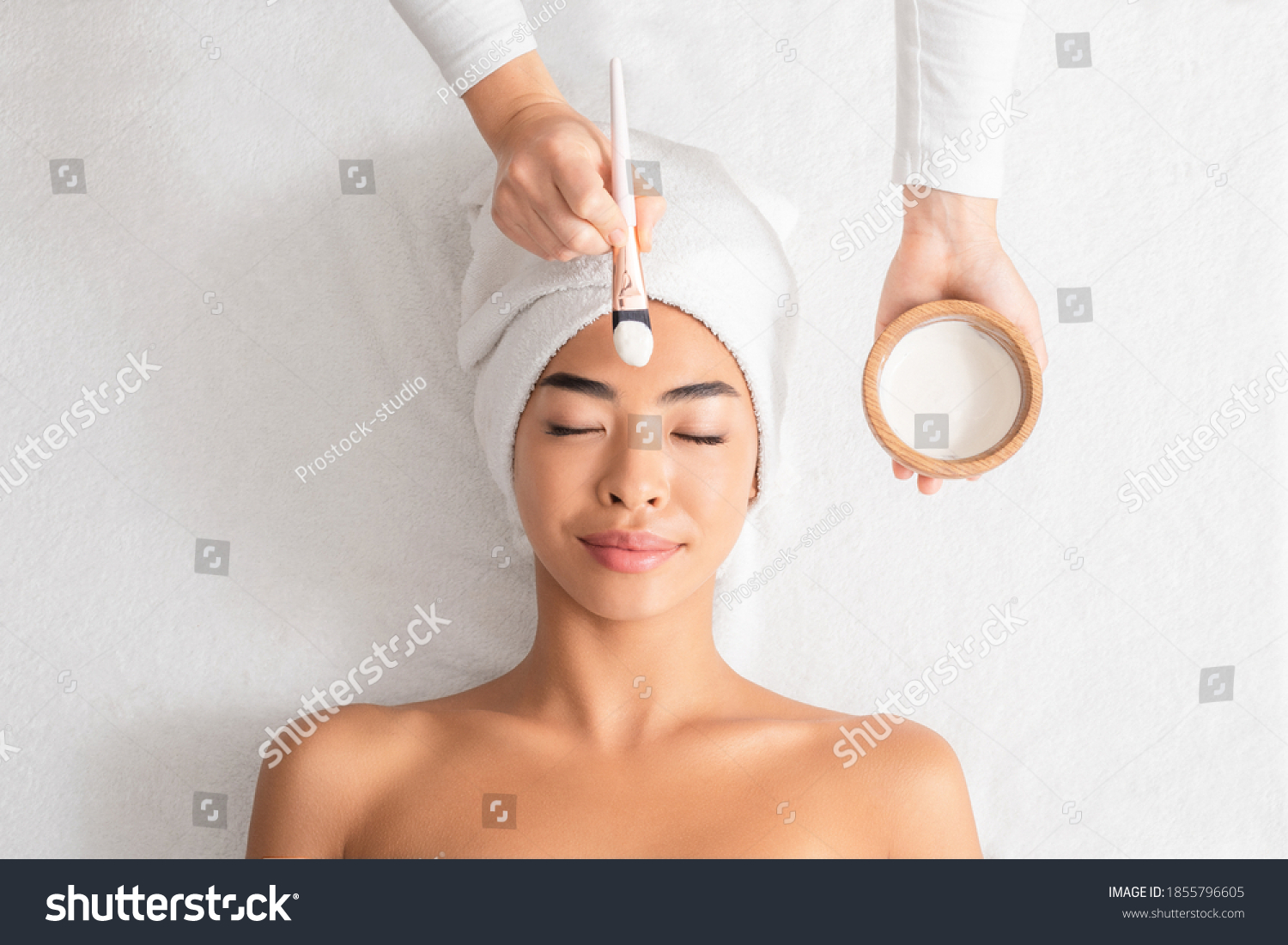 Beauty Treatments. Cosmetologist applying clay mask on asian woman face at spa salon  relaxed korean lady with towel on head enjoying wellness day at beauty center  top view with free space
