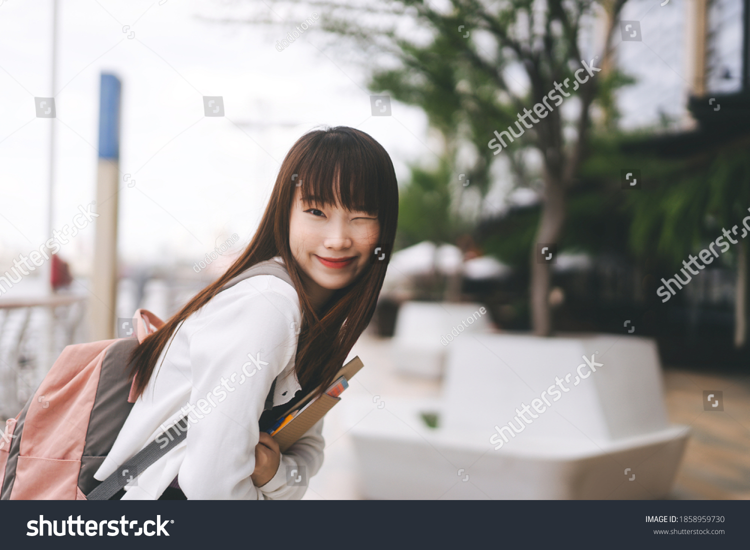 College people lifestyle on day time concept. Portrait of young adult asian student woman. Hold notebook and wear backpack at outdoor. City and river blur background.