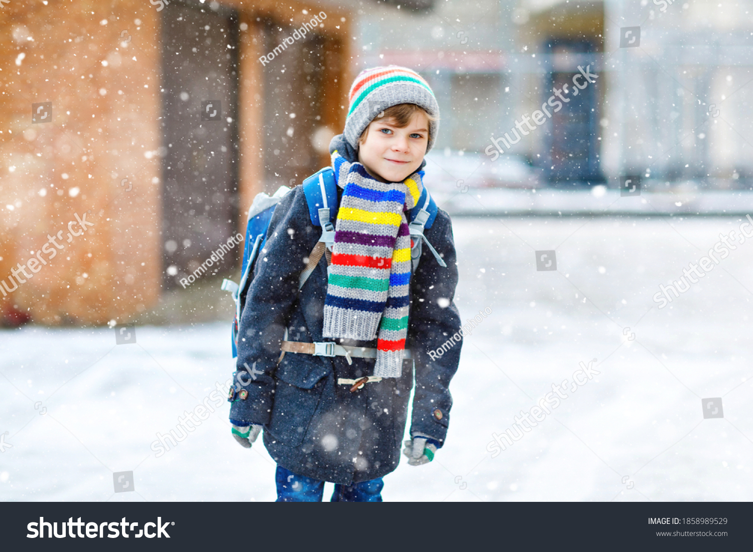 Little school kid boy of elementary class walking to school during snowfall. Happy child having fun and playing with first snow. Student with backpack or satchel in colorful winter clothes.