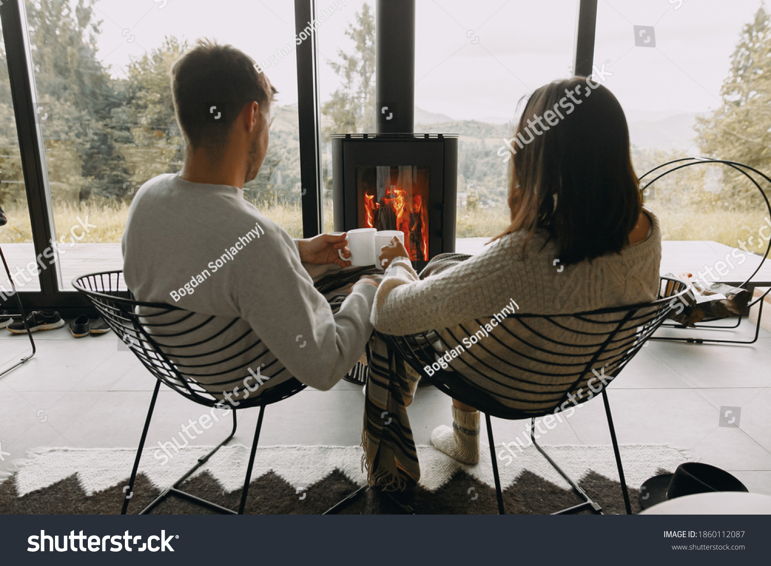 Stylish couple in sweaters with warm cups of tea relaxing at modern black fireplace with view on mountains. Cozy warm family moments at cold season. Young family on vacation