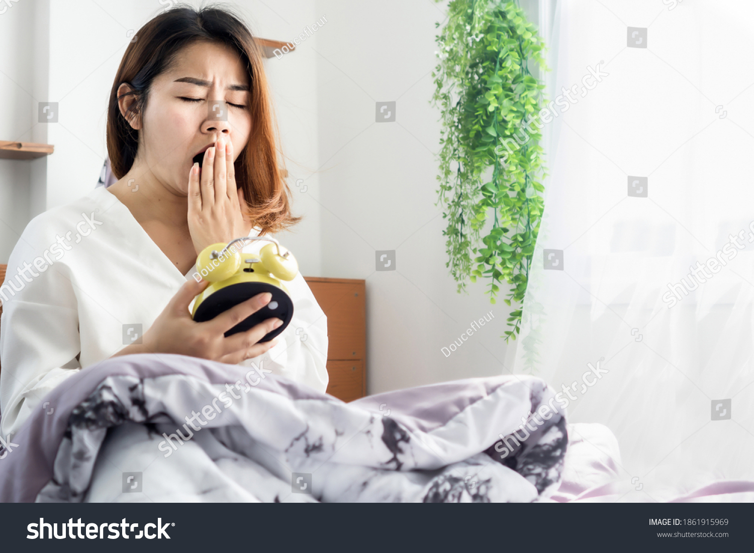 Asian woman yawning bed hand holding alarm clock feeling tired and sleepy 