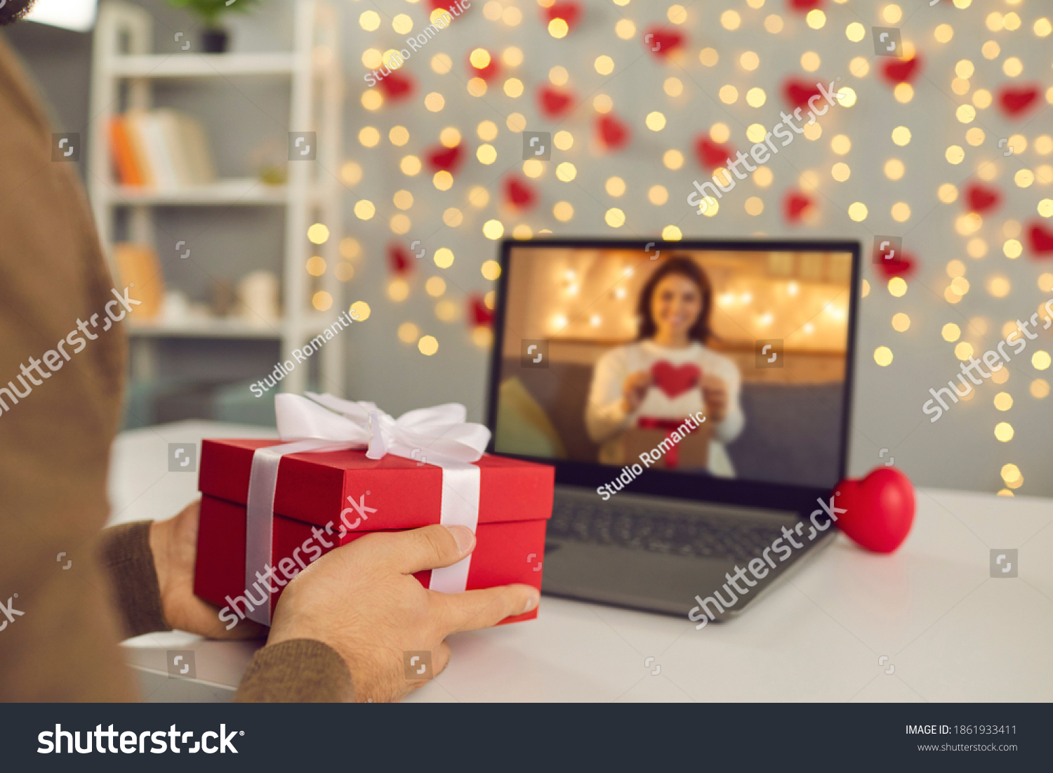 Video call on Saint Valentine's Day. Couple sending each other love and showing presents on virtual date. Closeup of man's hands holding gift box. Shimmering yellow lights soft focus selective focus