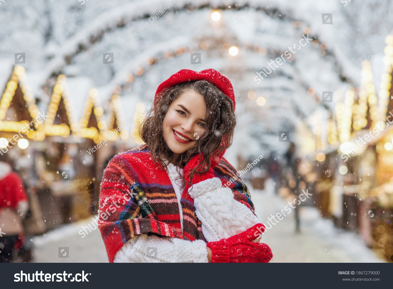 Winter  Christmas  New Year holidays concept: happy smiling woman posing at street festive market. Model wearing red beret  tartan scarf  red gloves  white knitted sweater. Copy  empty space for text