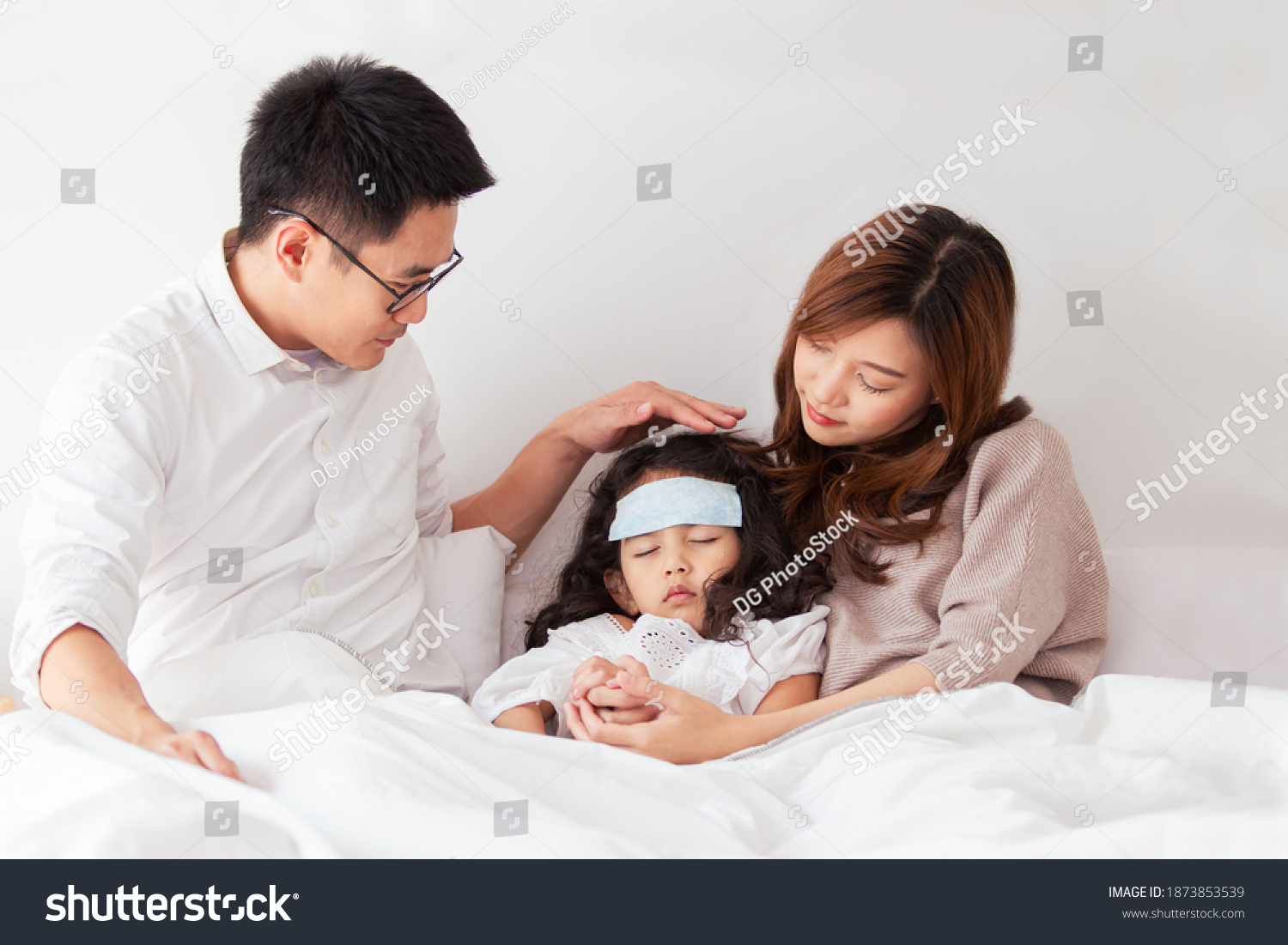 Asian sick girl sleeping on the bed with fever gel pack on her forehead. Father and mother take care of their little young daughter who got a fever from flu virus. sick little kid laying on the bed.