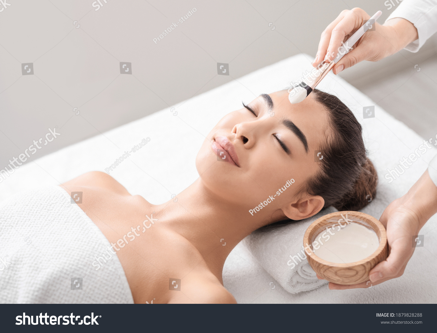 Beauty Treatment. Professional beautician applying clay mask to asian woman at spa salon  young attractive korean female lying on massage table  enjoying face care procedures  closeup shot  free space