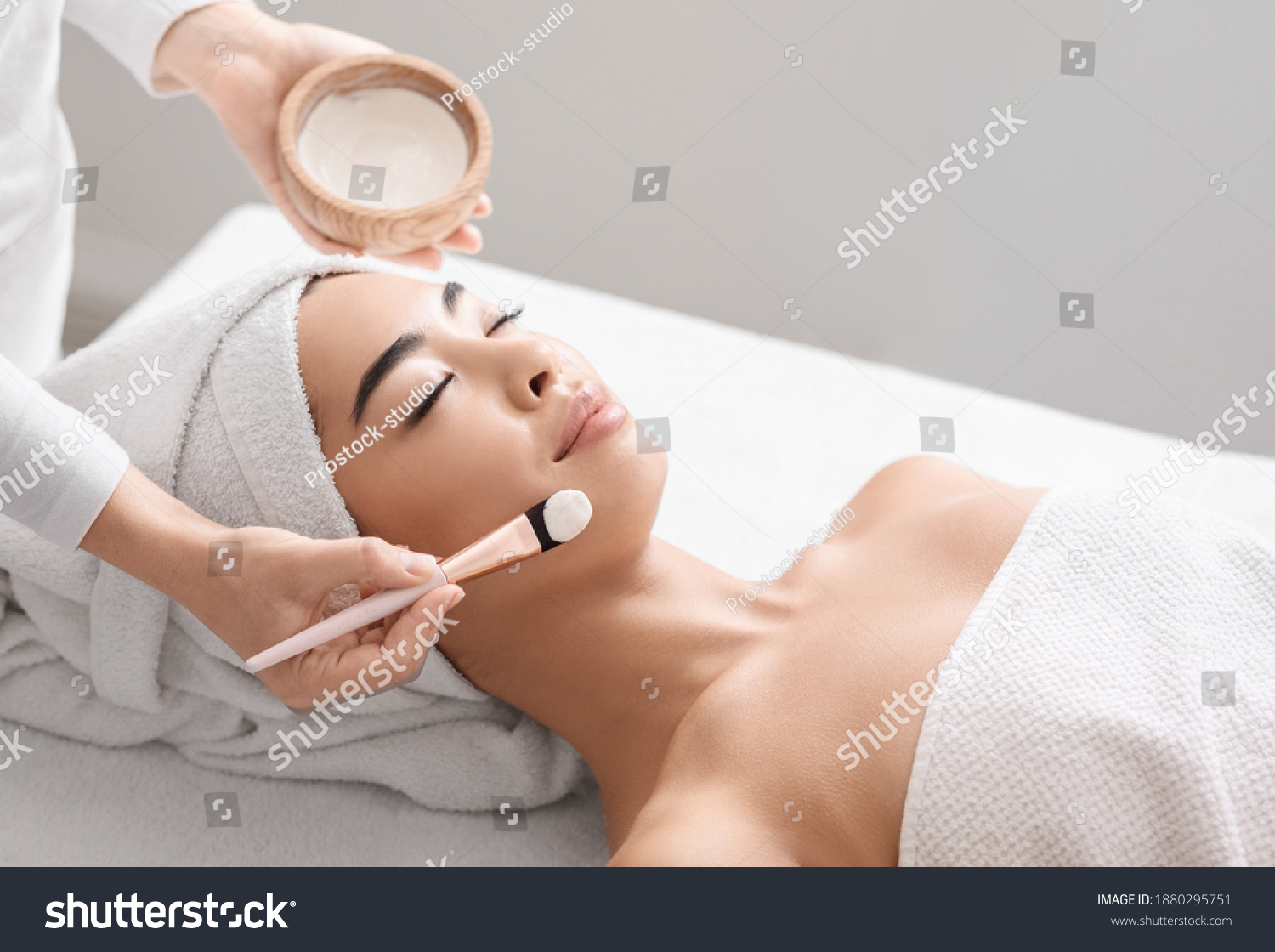 Attractive young asian woman receiving skin treatment at spa center. Professional cosmetologist doctor applying moisturising clay mask on beautiful korean lady's face closeup shot with copy space