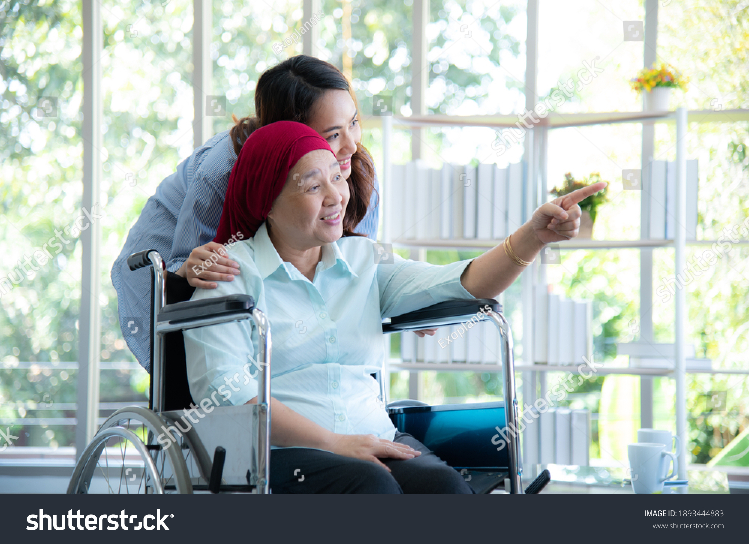 Asian elderly mother who sitting on wheelchair and wearing red headscarf  show her lovely daughter to look at something through window very happily and smile after she survive from cancer or leukemia.