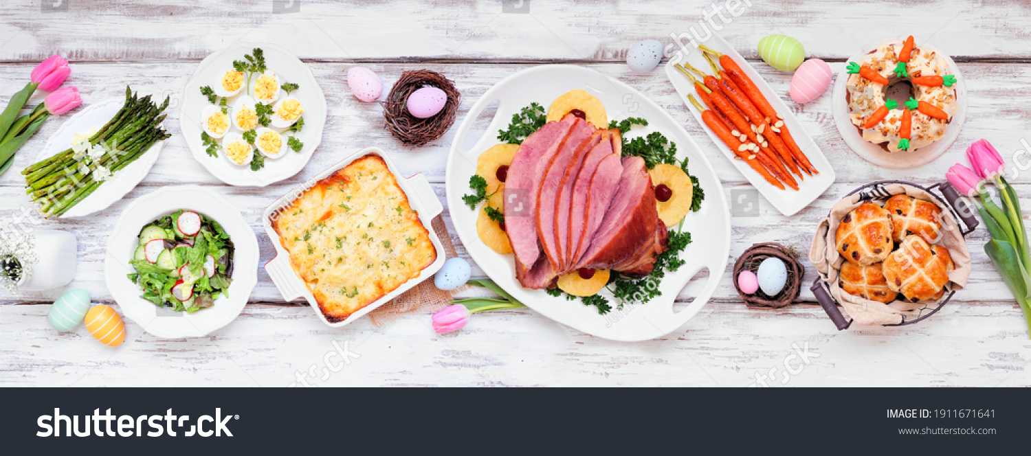 Classic Easter ham dinner. Top down view table scene on a white wood banner background. Ham scalloped potatoes eggs hot cross buns carrot cake and vegetables.
