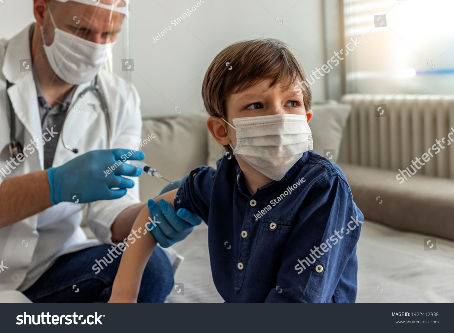 The smiling boy is looking away while his doctor holding a syringe next to his arm. Pediatrician makes vaccination to cute Caucasian boy. Boy in medical face mask getting flu shot by doctor at home.