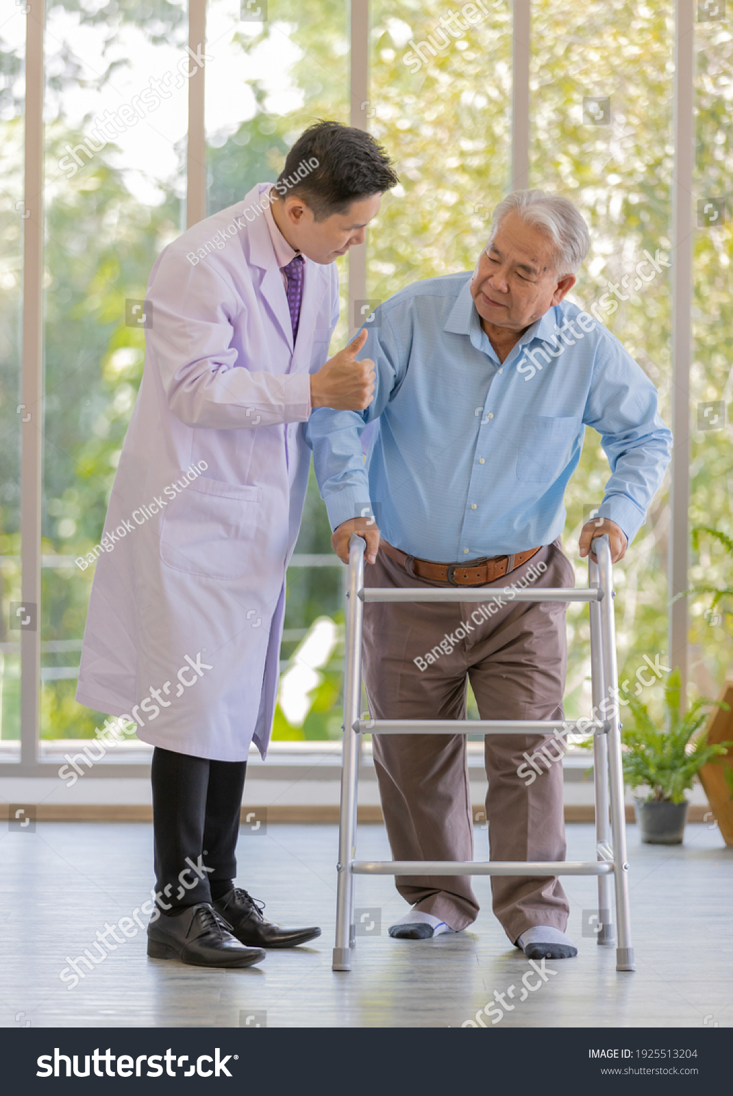 A young male asian doctor wearing white lab coat try to supporting his patient by holding his right arm when an old fat gray hair patient wearing light blue shirt and brown pants standing.