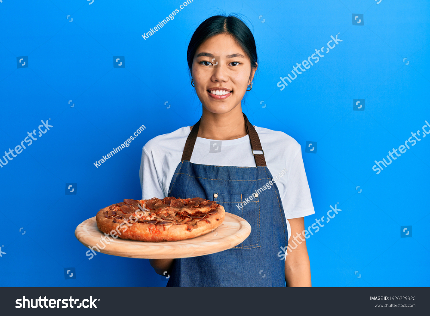 Young chinese woman wearing waiter apron holding italian pizza looking positive and happy standing and smiling with a confident smile showing teeth