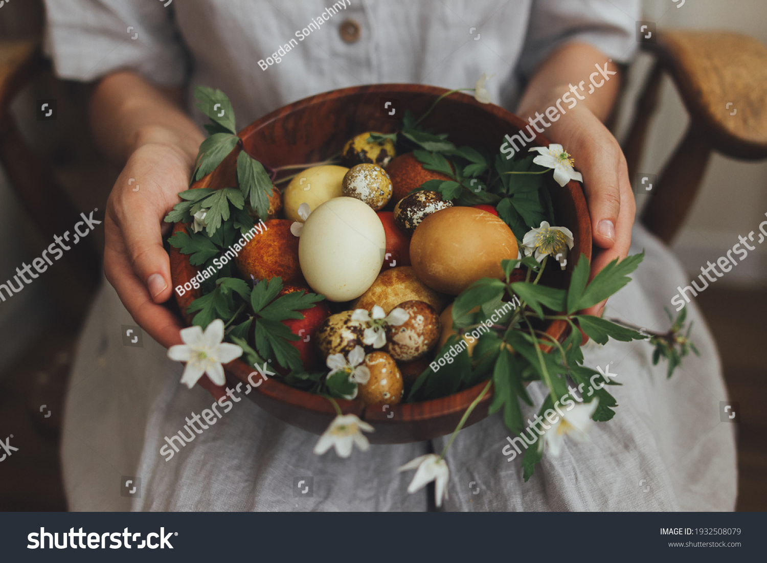 Easter eggs and spring flowers in wooden bowl in hands on background of woman in rustic linen dress. Stylish easter and quail eggs in natural dye and spring blooms. Aesthetic holiday