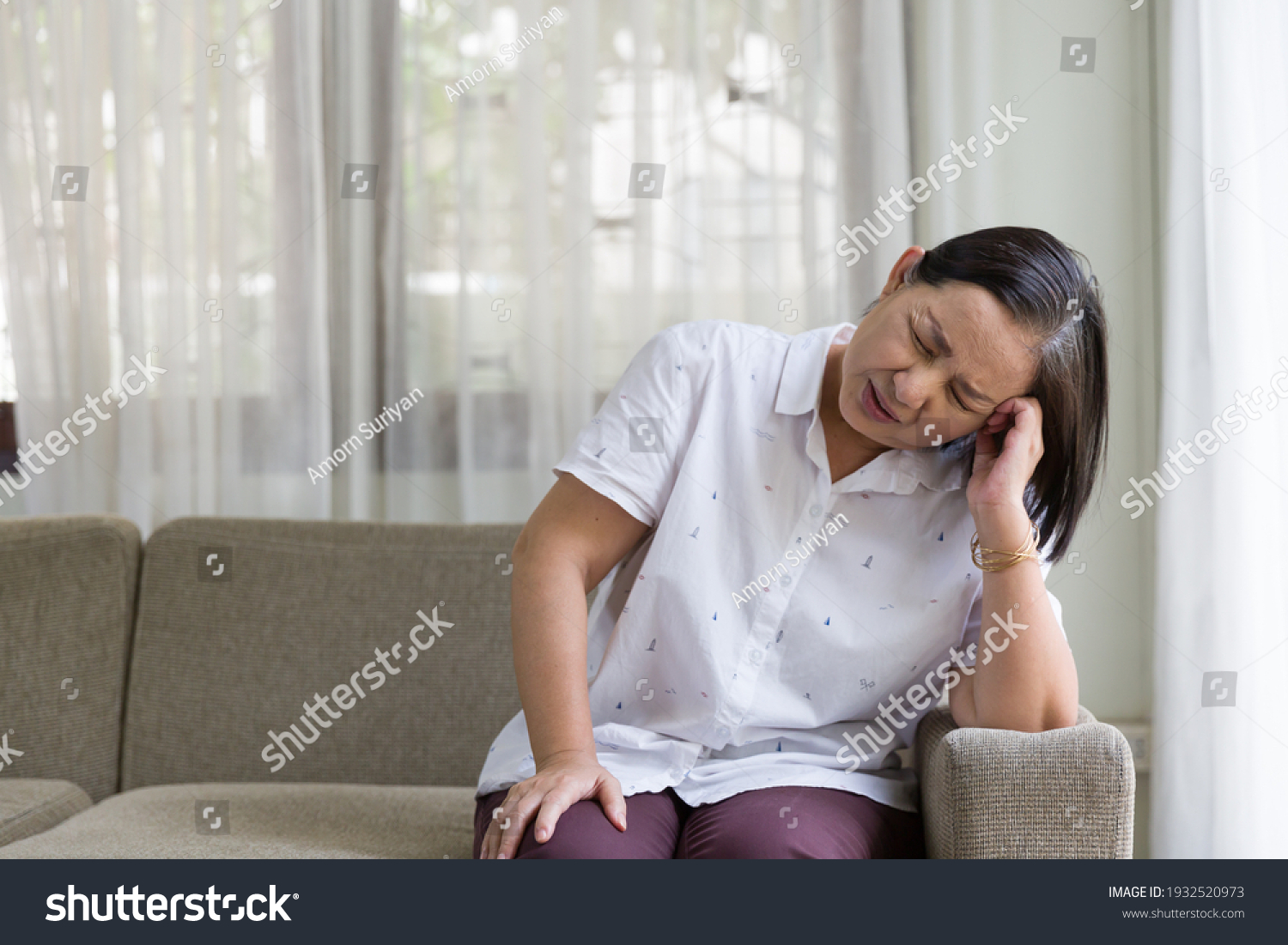 Elderly Asian woman sitting and having a headache and touching her head with her hands at home. Retirement quarantine and health care concept