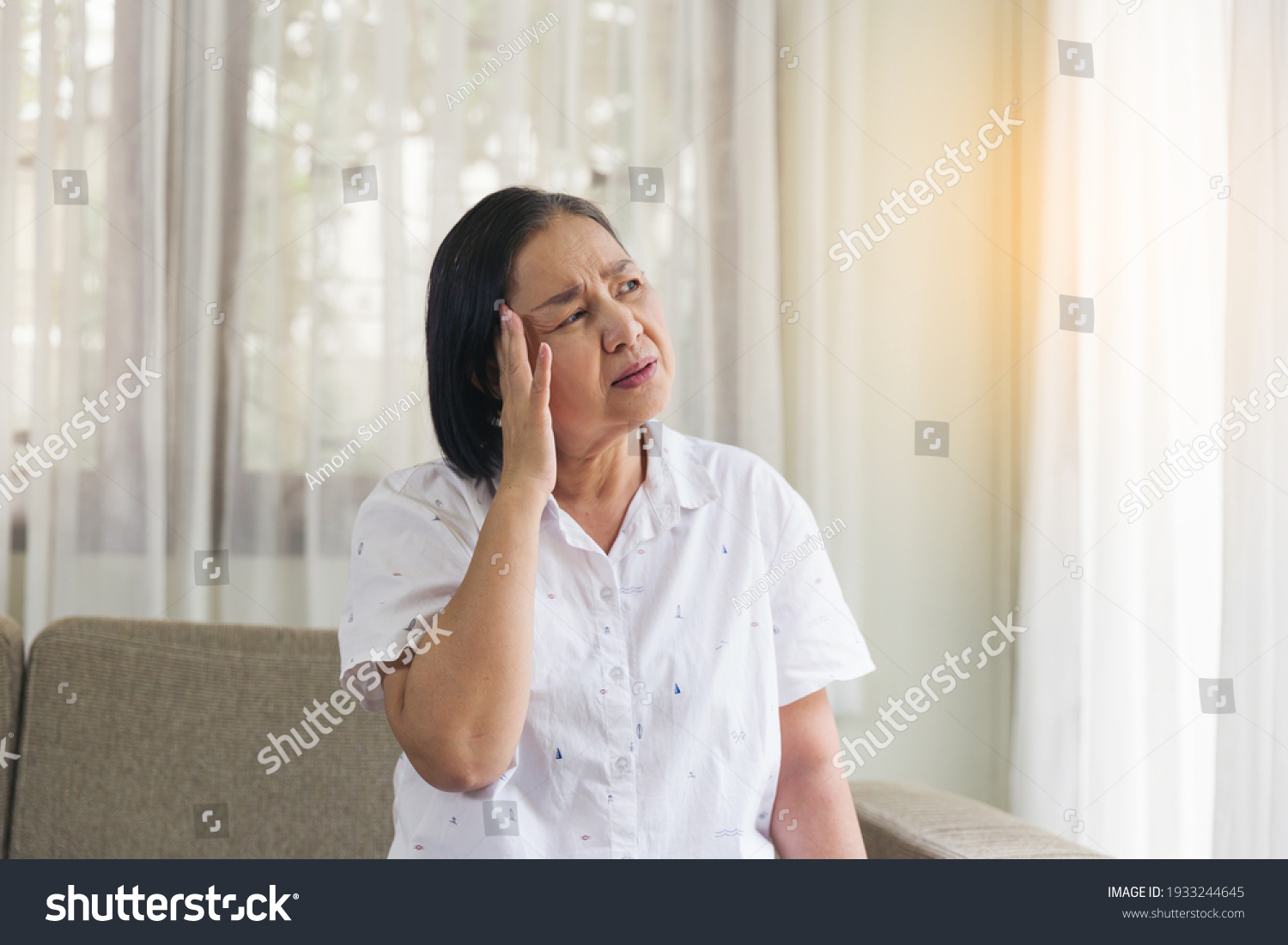 Elderly Asian woman sitting and having a headache and touching her head with her hands at home. Retirement quarantine and health care concept