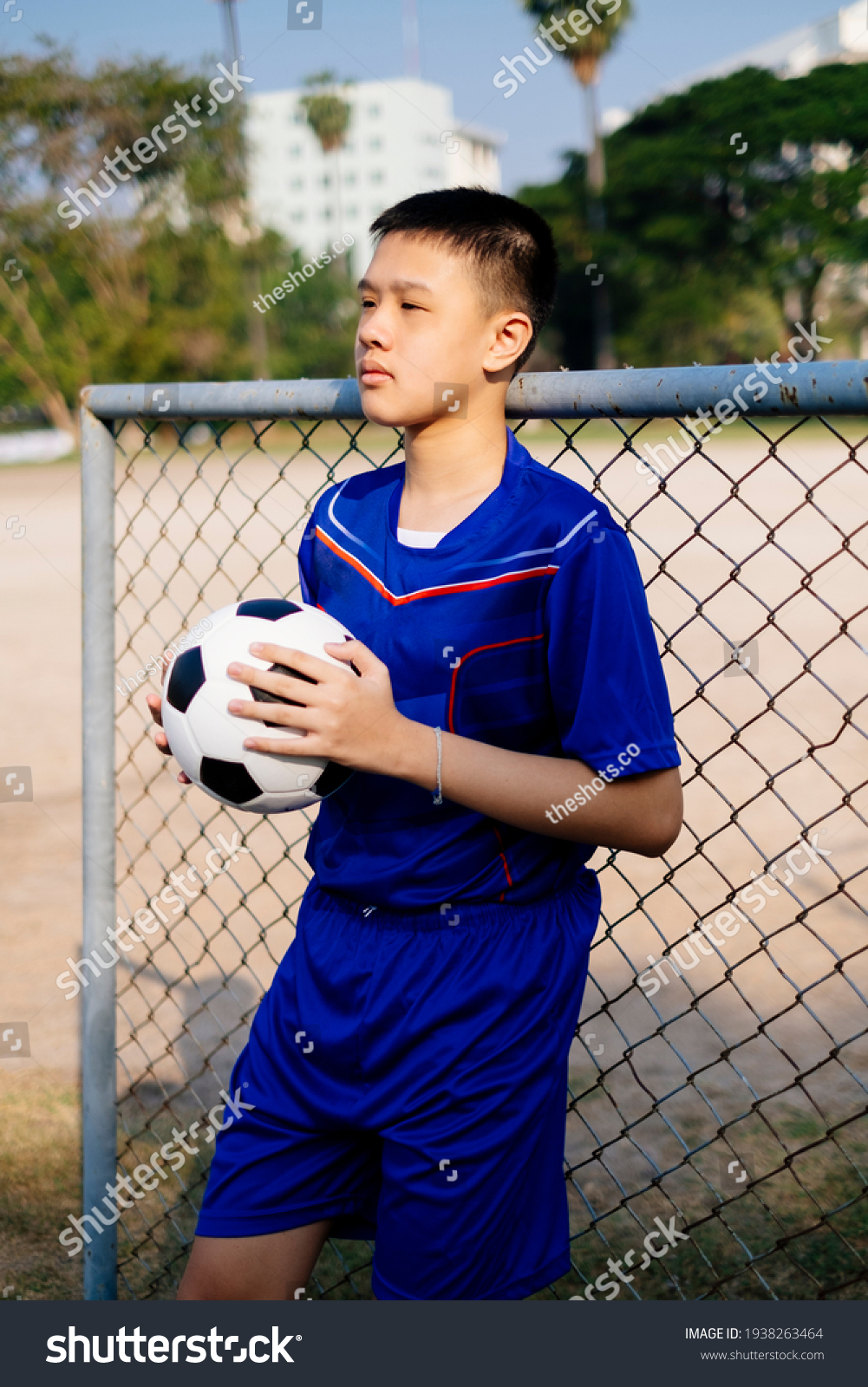 A boy in blue football suit standing and hold his ball at the fence.