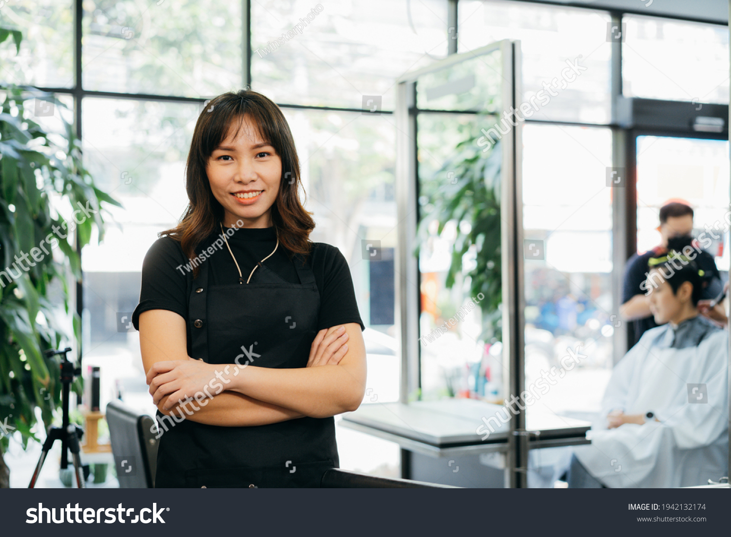 Portrait of happy successful young Asian salon owner standing wearing apron with folded hands smiling while looking at camera